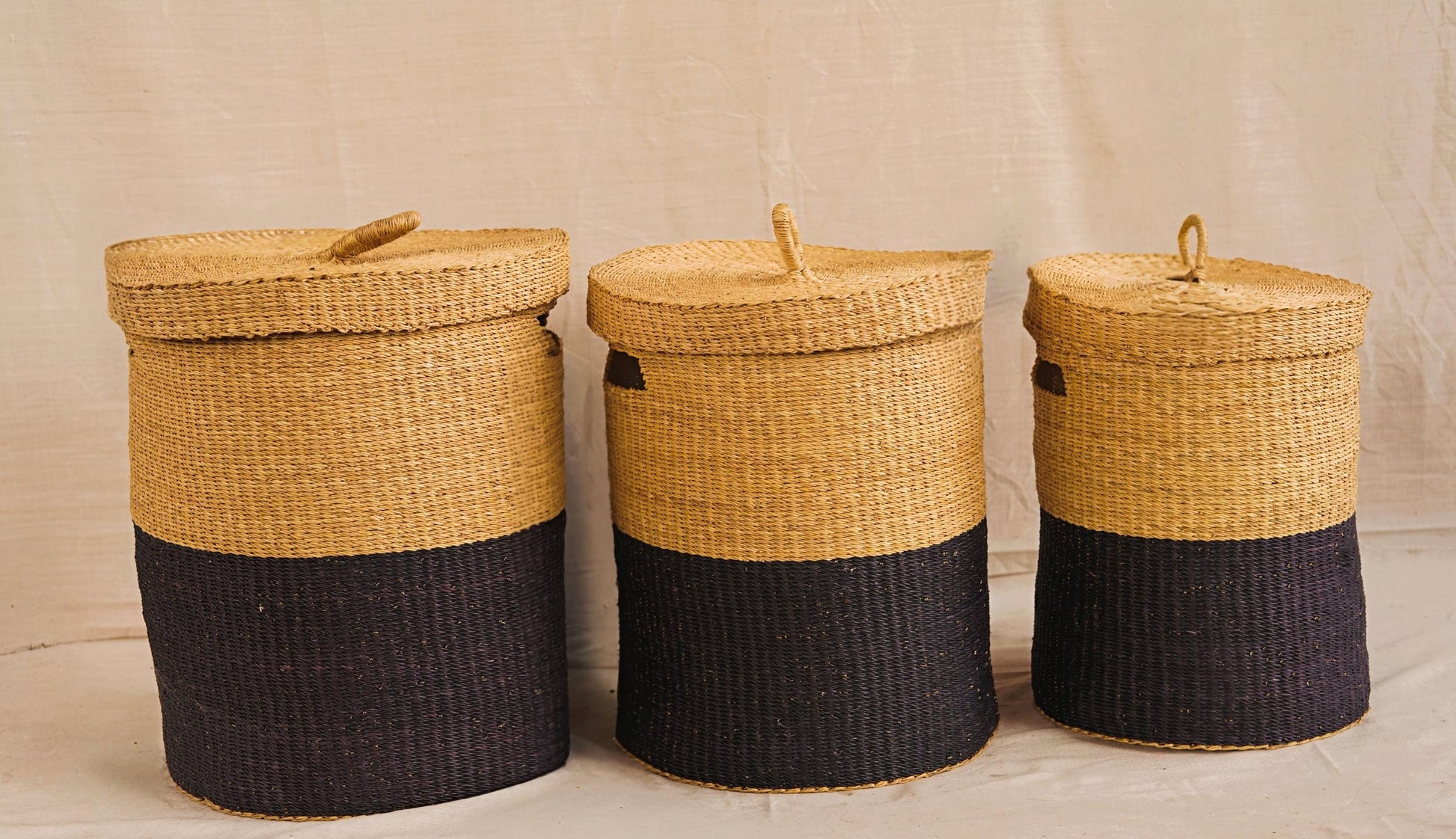 Three handwoven Bolga storage baskets in varying sizes with lids, featuring a natural and black color pattern, made from elephant grass