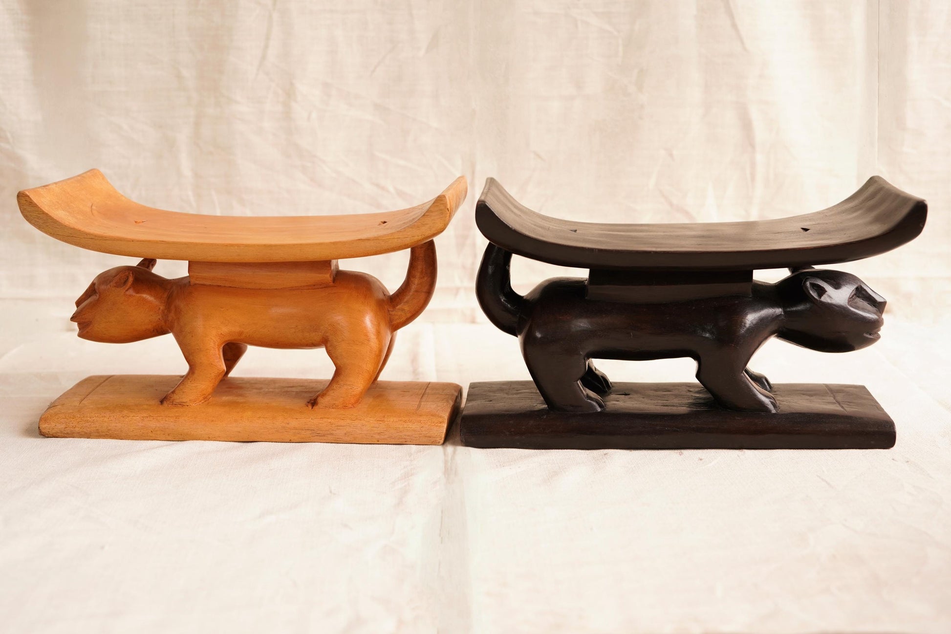 Two Ashanti hand-carved wooden stools with curved seats, one in natural wood color and the other stained in a darker shade, displayed against a white background.