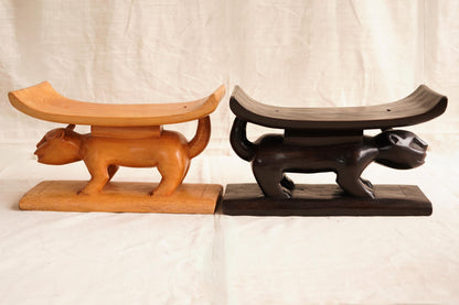 Two Ashanti hand-carved wooden stools with curved seats, one in natural wood color and the other stained in a darker shade, displayed against a white background.