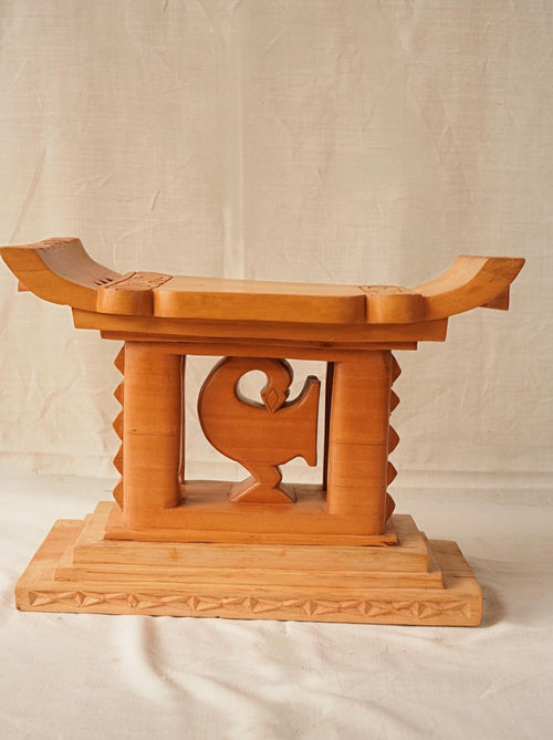 A hand-carved wooden Ashanti stool with a curved seat and arched base, featuring the Akan Adinkra symbol 'Sankofa' on the front.