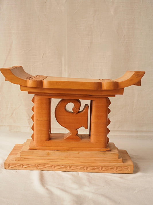 A hand-carved wooden Ashanti stool with a curved seat and arched base, featuring the Akan Adinkra symbol 'Sankofa' on the front.