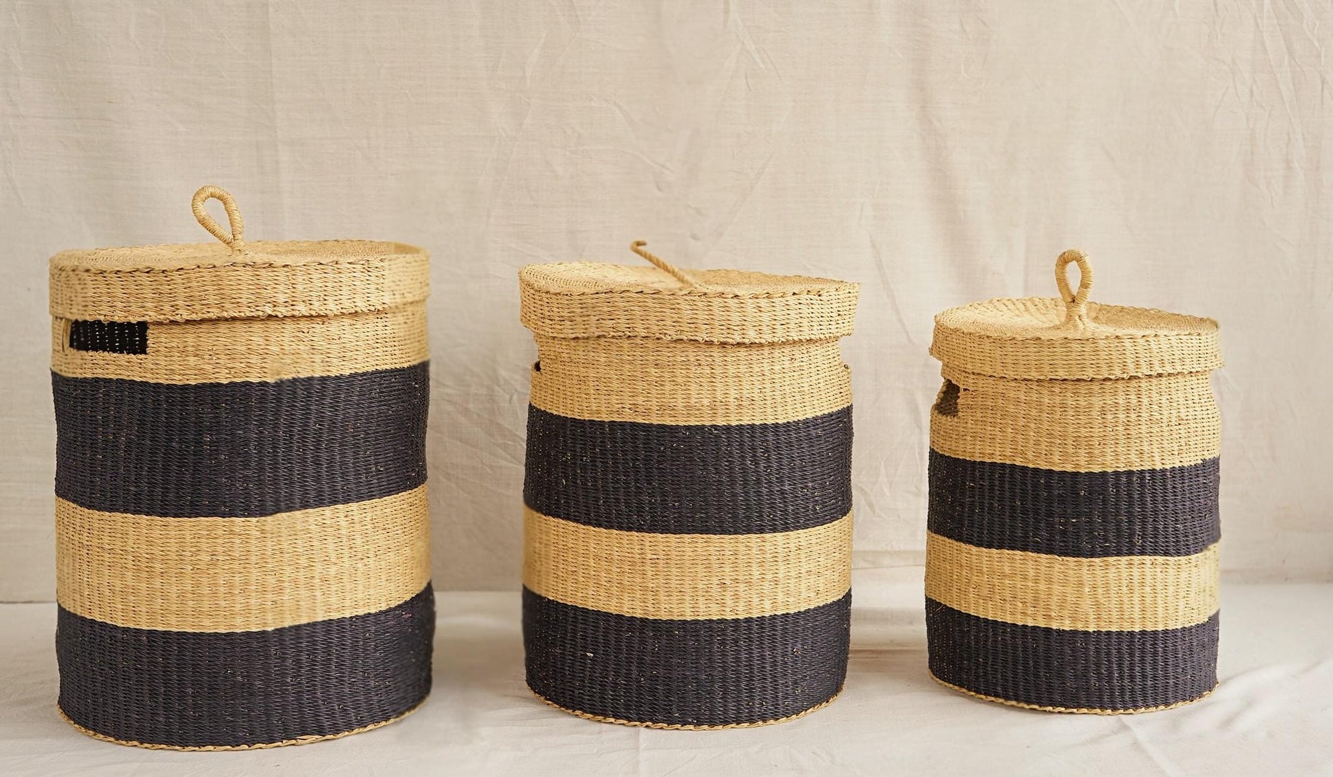Three handwoven Bolga storage baskets with lids, in varying sizes, featuring natural and black stripes and leather-wrapped handles.