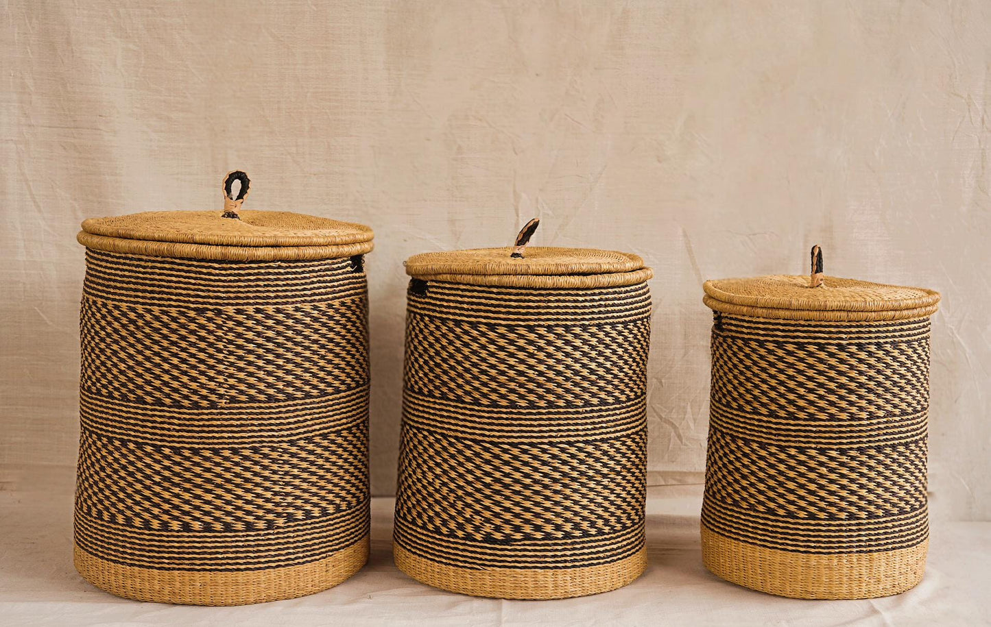 Three cylindrical Bolga storage baskets with lids, in different sizes, featuring a natural weave pattern and leather-wrapped handles.