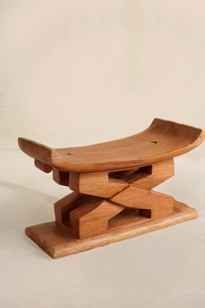 A miniature wooden Ashanti  stool with a curved seat and X-shaped base on a beige background
