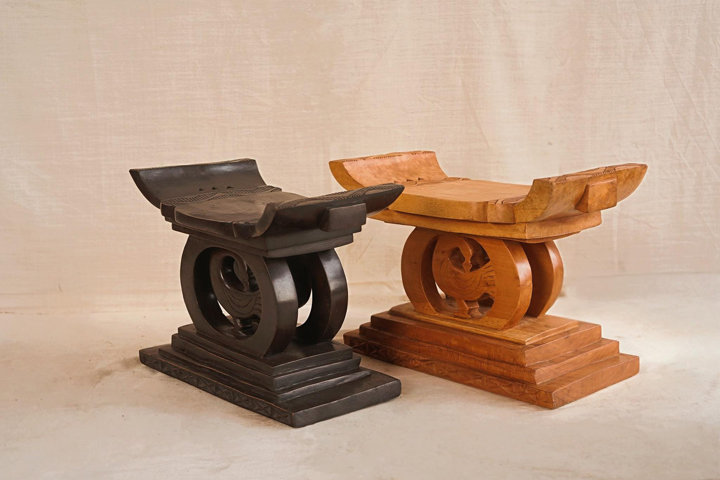 Two Ashanti hand-carved wooden stools with natural patina, one in dark brown and the other in light brown, displaying traditional curved seats and arched bases.